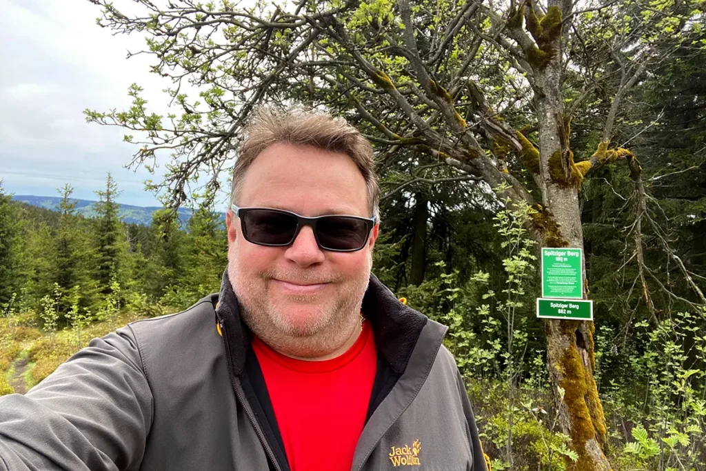 Selfie: Torsten Widder mit Sonnenbrille lächelt in Kamera bei Wanderung im Wald, im Hintergrund Schild Spitziger Berg